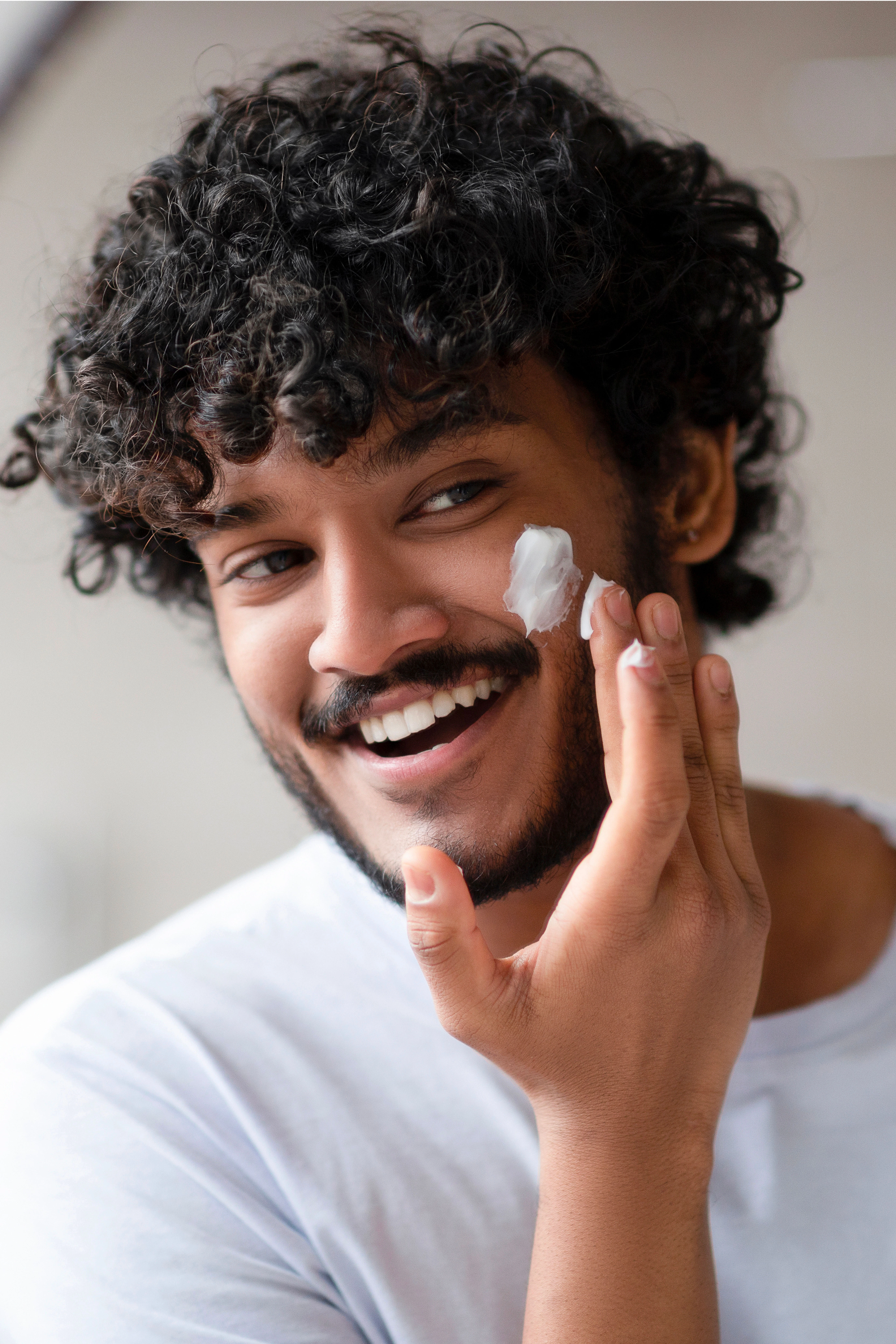 homme-jeune-creme Jeune homme appliquant soigneusement une crème hydratante sur son visage, dans une salle de bain bien éclairée.